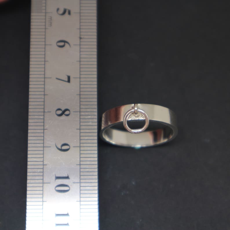 May include: A silver ring with a small circular charm attached. The ring is 8 mm wide and is measured against a ruler.