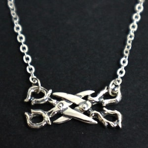 Könnte beinhalten: Eine silberfarbene Halskette mit einem Anhänger in Form einer Schere. Die Schere hat detaillierte, gebogene Griffe und Klingen, die an einer feinen Kette befestigt sind. Die Halskette wird vor einem dunklen Hintergrund gezeigt.