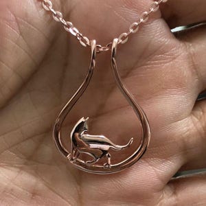 Puede incluir: Collar con colgante de gato en oro rosa. El colgante presenta un gato estilizado dentro de un marco en forma de lágrima. La cadena y el colgante son de color oro rosa a juego, creando un diseño cohesivo y elegante. El gato está representado de pie.