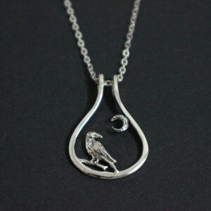 May include: A silver pendant necklace. The pendant has a teardrop shape, with a bird on a branch and a crescent moon with a gem inside. The necklace hangs from a delicate chain. The pendant is approximately 3 cm in height.
