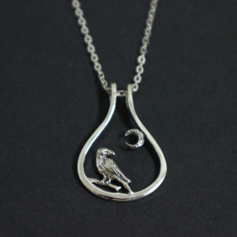 May include: A silver pendant necklace. The pendant has a teardrop shape, with a bird on a branch and a crescent moon with a gem inside. The necklace hangs from a delicate chain. The pendant is approximately 3 cm in height.