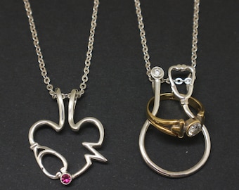 May include: Two silver necklaces with pendants. One necklace features a heart-shaped pendant with a stethoscope design and a small pink gemstone. The other necklace features a stethoscope pendant with a ring and a small clear gemstone.