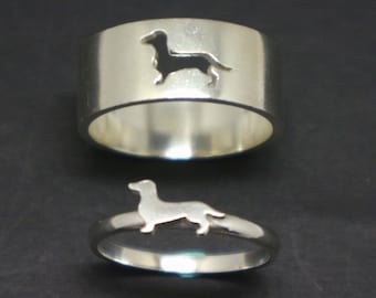 May include: Two silver rings with a dachshund silhouette cutout. The larger ring is a band with the dachshund cutout on the top. The smaller ring is a thin band with the dachshund cutout on the side.