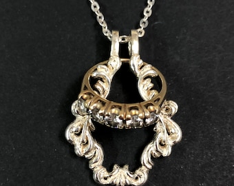 May include: A silver necklace with a pendant featuring a ring with a row of clear stones set in a gold band. The ring is suspended within a silver frame with ornate scrollwork.