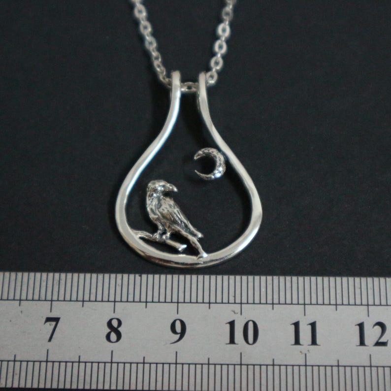 May include: A silver pendant necklace featuring a bird perched on a branch inside a teardrop-shaped frame. A crescent moon is also inside the frame. The necklace is on a silver chain.