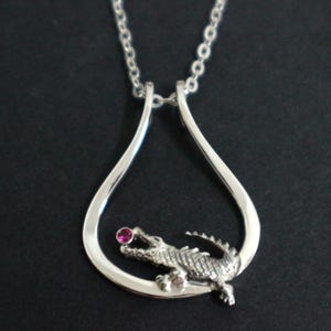 May include: Silver pendant necklace with a detailed alligator and a small, round, pink gemstone. The alligator sits within a teardrop-shaped frame, suspended from a fine chain. The necklace is shown against a dark backdrop.