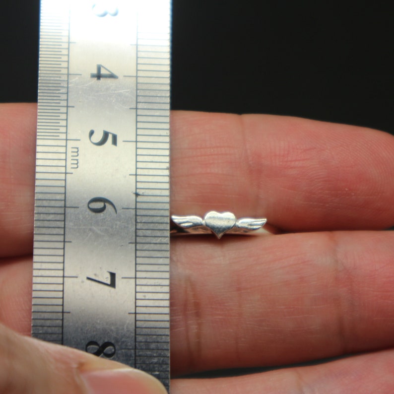 May include: A silver charm with a heart design and wings. The charm is about 6 mm wide and is measured against a ruler.