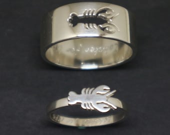 May include: Two silver rings with lobster designs. One ring is wide with a lobster cutout, the other is a thin band with a lobster accent. Both rings have engraved text. The rings are displayed on a black background.