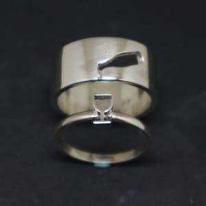 May include: Two silver wedding bands with a wine bottle and wine glass design. The bands are stacked on top of each other.