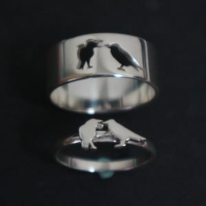 May include: Two silver-toned rings featuring a cutout design of two birds facing each other. The wider band displays the birds as a negative space silhouette, while the thinner band features the birds as a raised metal cutout on the top.