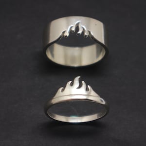 Pode incluir: Duas alianças de prata com um design de chama. A aliança superior tem um design de chama recortado, enquanto a aliança inferior tem um design de chama sólido.