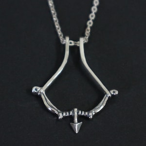 May include: A silver-toned pendant necklace featuring a bow and arrow design. The pendant has a curved bow shape with an arrow pointing downwards. The necklace chain is a delicate link style. The background is a solid black.