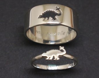 May include: Two silver rings featuring a dinosaur design. The larger ring has a cutout of a dinosaur and the inscription "my puzzle fits". The smaller ring has a solid dinosaur design.