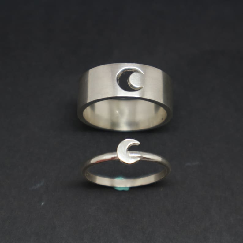 May include: Two silver rings with a crescent moon design. The larger ring has a cut-out crescent moon, while the smaller ring has a solid crescent moon.