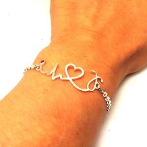 Peut inclure: Bracelet argenté avec un motif de battement de cœur, un cœur et un symbole de stéthoscope. Une petite pierre rouge orne le mot "i". Le bracelet est relié par une fine chaîne.