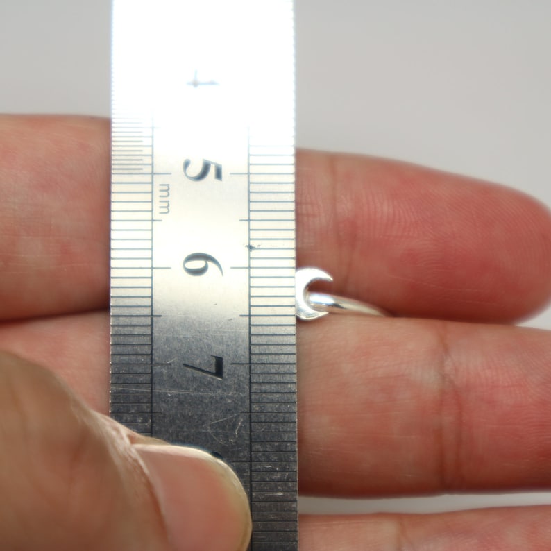 May include: A silver ring with a crescent moon design is being measured with a ruler. The ring is on a finger and the ruler shows a measurement of 6.7 mm.
