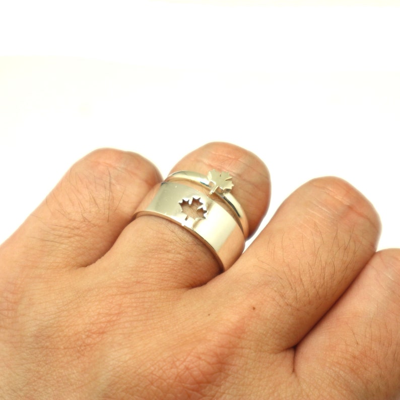 May include: A set of two silver rings. One ring has a cutout maple leaf design, and the other ring has a small maple leaf on top. The rings are worn on a finger.