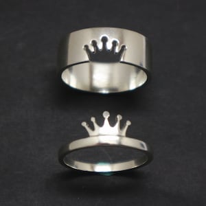 Może przedstawiać: Dwa srebrne pierścionki z wzorem korony. Górny pierścionek ma koronę wyciętą z obrączki. Dolny pierścionek ma koronę na górze obrączki.