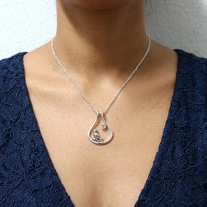 May include: Silver pendant necklace with a bird on a branch inside a teardrop frame, with a crescent moon. The necklace is worn over a dark blue top. The chain is visible.