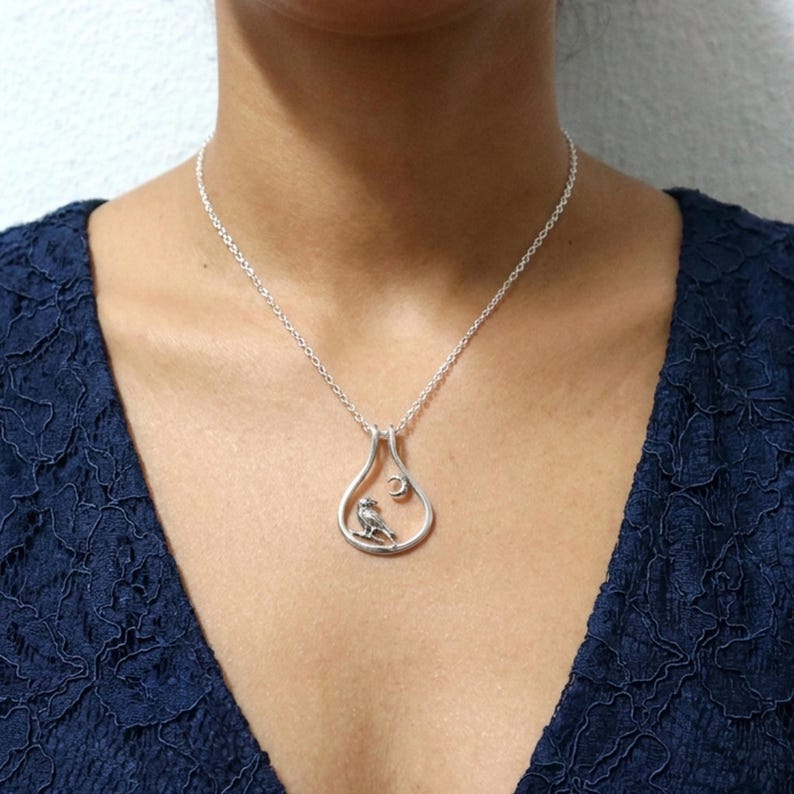 May include: Silver pendant necklace with a bird on a branch inside a teardrop frame, with a crescent moon. The necklace is worn over a dark blue top. The chain is visible.