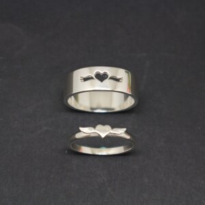 May include: Two silver rings with heart cutouts. The larger ring has a heart with an arrow through it. The smaller ring has a heart with wings.