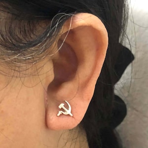 Puede incluir: Primer plano de la oreja de una persona con un pequeño pendiente de plata con un diseño de martillo y hoz.