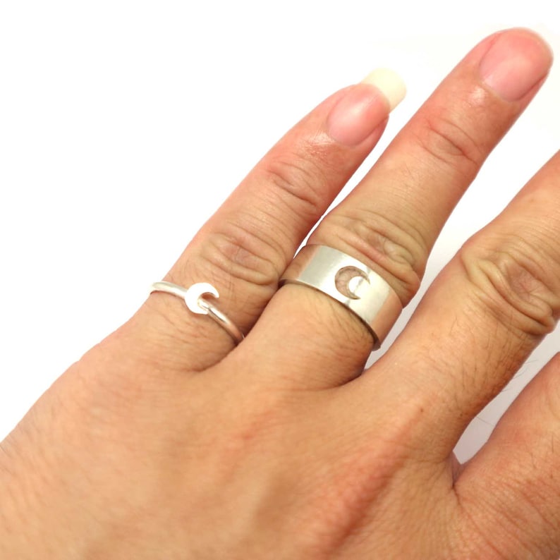 May include: Two silver rings with crescent moon designs. The ring on the left is a thin band with a small crescent moon. The ring on the right is a thicker band with a larger crescent moon cut out of the band.