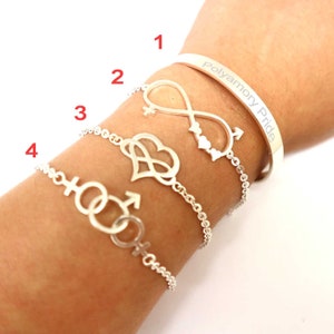 Puede incluir: Un conjunto de pulseras de plata con cuatro dijes diferentes. Los dijes incluyen un símbolo de infinito con un corazón y una flecha, un corazón con un símbolo de infinito y dos círculos entrelazados con una flecha y un símbolo para femenino y masculino. La pulsera también incluye una pulsera de plata con el texto "Polyamory Pride".
