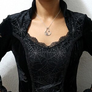 May include: A silver pendant necklace with a teardrop-shaped charm. The charm features a small, detailed design. The person is wearing a black velvet dress with spiderweb detailing and a lace neckline.
