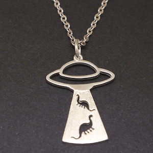 May include: Silver-toned necklace featuring a pendant shaped like a UFO with a beam of light. Inside the beam are two dinosaur silhouettes. The necklace has a delicate chain and is set against a dark background.