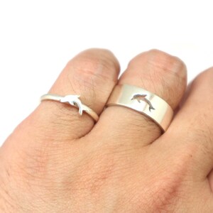 May include: Two silver rings with a dolphin design. The ring on the left has a solid dolphin, while the ring on the right has a cut-out dolphin.