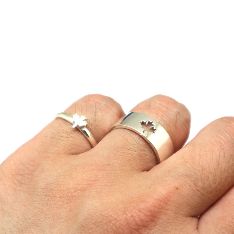 May include: Two silver rings, one a band with a maple leaf cutout, the other a delicate ring with a maple leaf design. The rings are displayed on a hand against a white background. The rings are likely made of sterling silver.