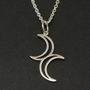 Può includere: Collana con pendente argentato a forma di luna crescente stilizzata. Il pendente è sospeso a una catenina sottile. La collana è mostrata su uno sfondo scuro, evidenziando la forma del pendente.