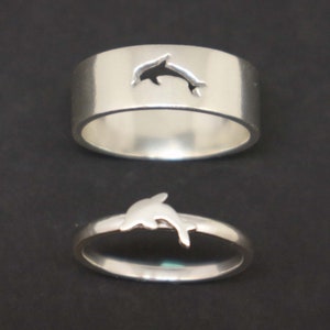 May include: Two silver rings with a dolphin cutout design. The top ring has a dolphin cutout, while the bottom ring has a solid dolphin design.