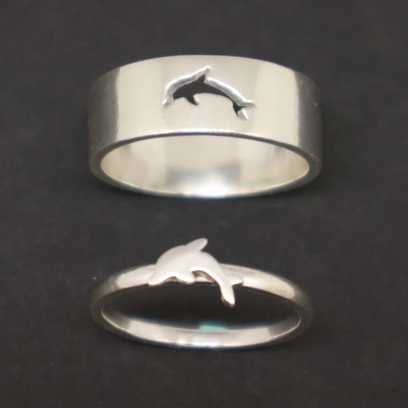 May include: Two silver rings with a dolphin cutout design. The top ring has a dolphin cutout, while the bottom ring has a solid dolphin design.