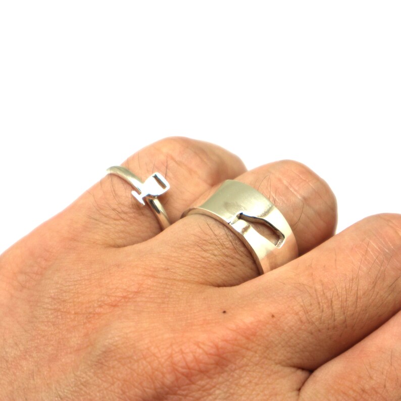 May include: Two silver rings on a hand. One ring is a simple band with a cutout design. The other ring is a thin band with a small, raised design.