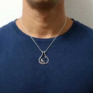 May include: A silver pendant necklace with a bird and crescent moon design. The teardrop-shaped pendant hangs from a fine chain. The person in the image is wearing a blue t-shirt. The necklace is a piece of jewellery.