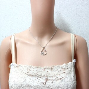 May include: A silver pendant necklace with a teardrop-shaped design. The pendant features a small bird and a clear stone. The necklace is worn by a person wearing a white lace top.