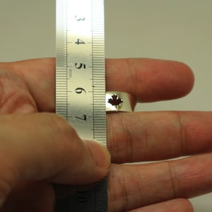 May include: A silver ring with a cut-out maple leaf design. The ring is being measured with a ruler. The ring's design features a small, dark red maple leaf. The ring is approximately 0.75 inches in diameter.