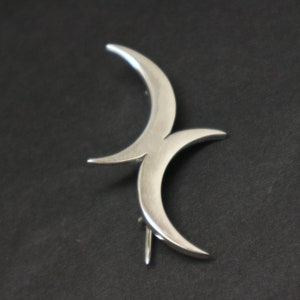 Pode incluir: Um broche de prata em forma de lua crescente com duas luas crescentes entrelaçadas.