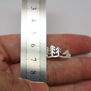May include: A silver ring featuring a cut-out design of a mountain range and pine trees. The ring is being measured with a ruler, showing it is approximately 7 cm in diameter.