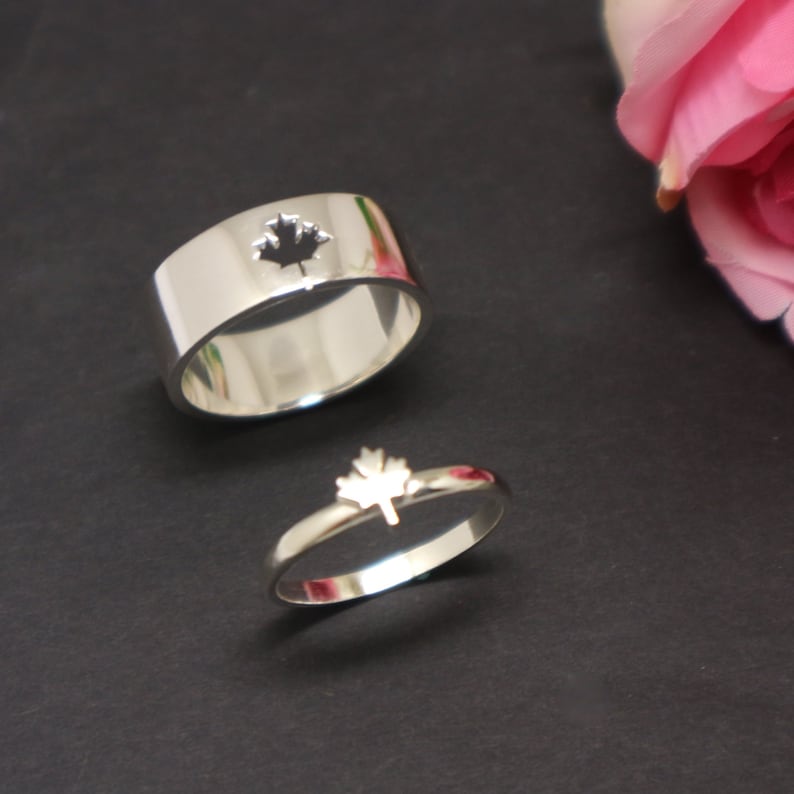 May include: Two silver rings with a maple leaf design. One ring is a wide band with a maple leaf cutout. The other ring is a thin band with a maple leaf on top. The rings are on a black surface.