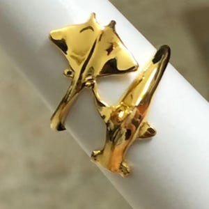 May include: A gold-colored ring featuring a stylized design of two manta rays. The ring is wrapped around a white cylindrical object, showcasing the intricate details of the marine life-inspired jewelry.