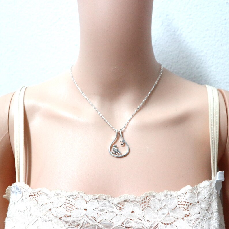 May include: A silver pendant necklace with a teardrop-shaped pendant, featuring a small bird and a crystal. The necklace is shown on a person wearing a white lace top.