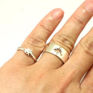 May include: Two silver rings, one thin with a dinosaur silhouette and one wider band with a dinosaur cutout. The rings are on a hand against a white background. The dinosaur design is visible on both rings.