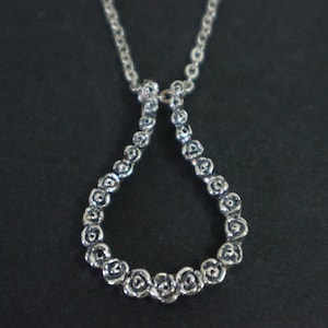 Puede incluir: Collar con colgante plateado con diseño de lágrima. El colgante está compuesto por pequeños elementos en forma de rosa. La cadena es de estilo eslabón fino, y el fondo es negro liso.