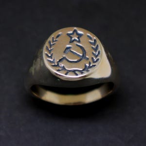 May include: A gold signet ring featuring a raised design of a hammer and sickle with a star, encircled by a laurel wreath.