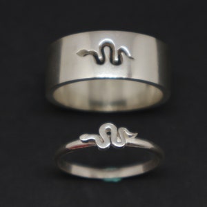 Puede incluir: Dos anillos de plata con un diseño de serpiente. El anillo más grande tiene un corte de serpiente en la parte superior, mientras que el anillo más pequeño tiene un diseño de serpiente que envuelve la banda.