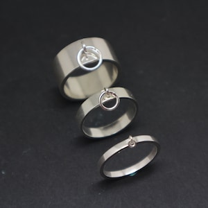 May include: Three silver rings with a small circle charm hanging from each. The rings are stacked on top of each other, with the largest ring at the bottom and the smallest ring at the top.