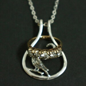 May include: Silver pendant necklace with a bird on a branch inside a teardrop frame. A gold ring with clear stones and a gold band is suspended within the frame. The necklace has a silver chain.